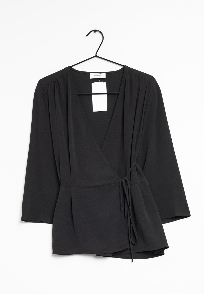 Blouse noire à manches longues avec col en V et ceinture latérale, suspendue à un cintre noir sur un fond blanc.