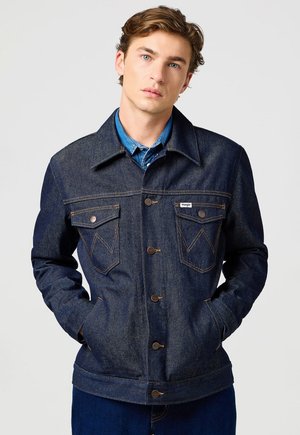 Młody mężczyzna o jasnobrązowych włosach, ubrany w ciemnoniebieską kurtkę dżinsową na niebieskiej koszuli, stoi z rękami w kieszeniach na jednolitym tle.