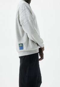 Sweatshirt gris avec un col rond, doté d'accents cousus et d'un logo sur le côté. Comprend un patch en étiquette verte en bas.