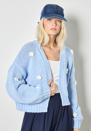 Hellblauer Strickcardigan mit Puffärmeln und bestickten weißen Blumen. Kombiniert mit einem schlichten weißen Oberteil und einer Jeanskappe.