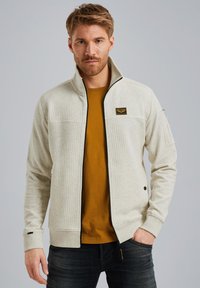 Veste zippée gris clair avec une partie avant texturée et des manches unies. Dotée d'une bordure noire, d'un patch logo et de poches latérales. Portée sur une chemise moutarde.