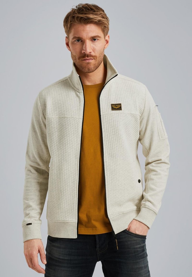 Veste zippée gris clair avec une partie avant texturée et des manches unies. Dotée d'une bordure noire, d'un patch logo et de poches latérales. Portée sur une chemise moutarde.