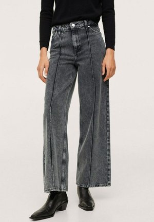 Kobieta ubrana w czarne, szerokie, wysokowalne jeansy z czarnymi botkami do kostki oraz czarną bluzkę z długim rękawem, stojąca na jednolitym tle.