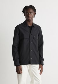 Camisa negra de botones con dos bolsillos en el pecho, diseñada con un corte recto, hecha de tela suave, con un sutil detalle del logo.