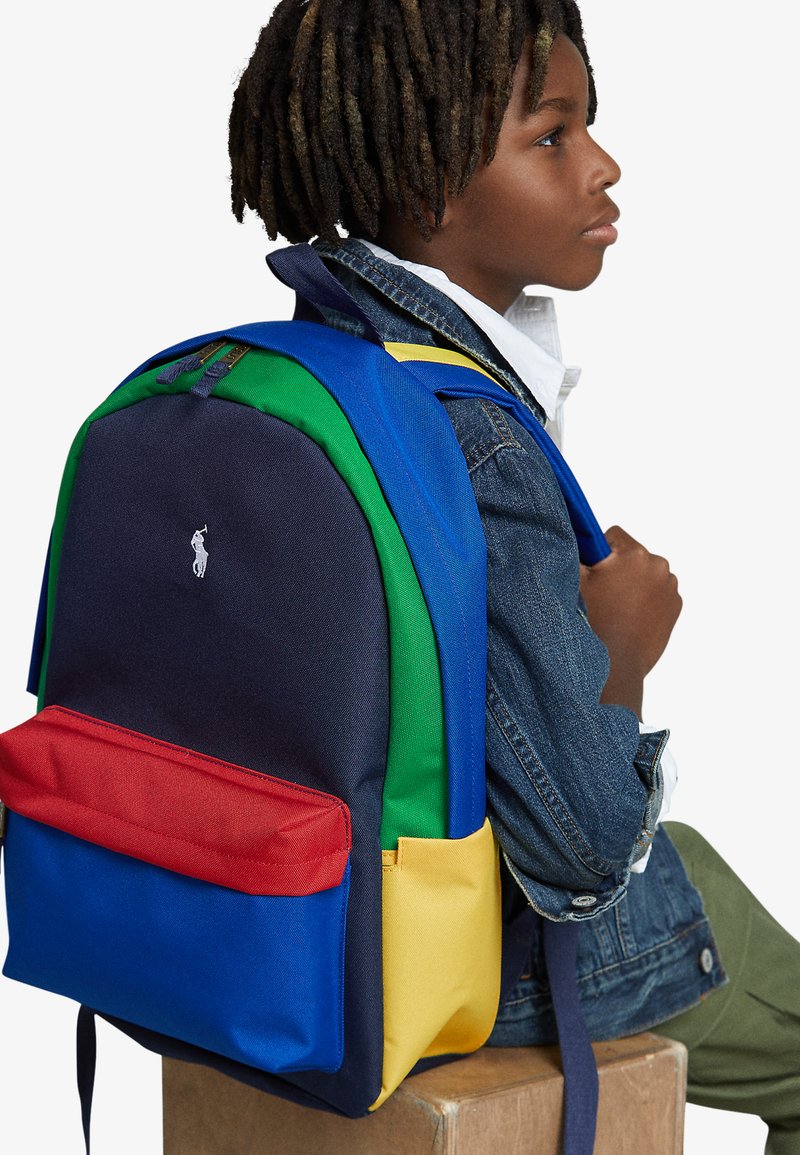 Mochila multicolor con paneles azul, verde, rojo y amarillo. Presenta un asa en la parte superior, correas ajustables y un pequeño logotipo en la parte frontal.