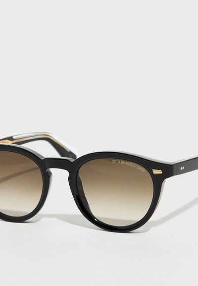 Μαύρα στρογγυλά γυαλιά ηλίου με καφέ βαθμιδωτούς φακούς και χρυσές λεπτομέρειες, τοποθετημένα σε λευκή επιφάνεια με κλίση.