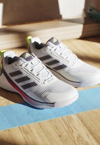 Baskets de sport blanches avec des accents gris et un design à trois rayures. Elles présentent un dessus texturé, des lacets et une boucle de talon verte vibrante.