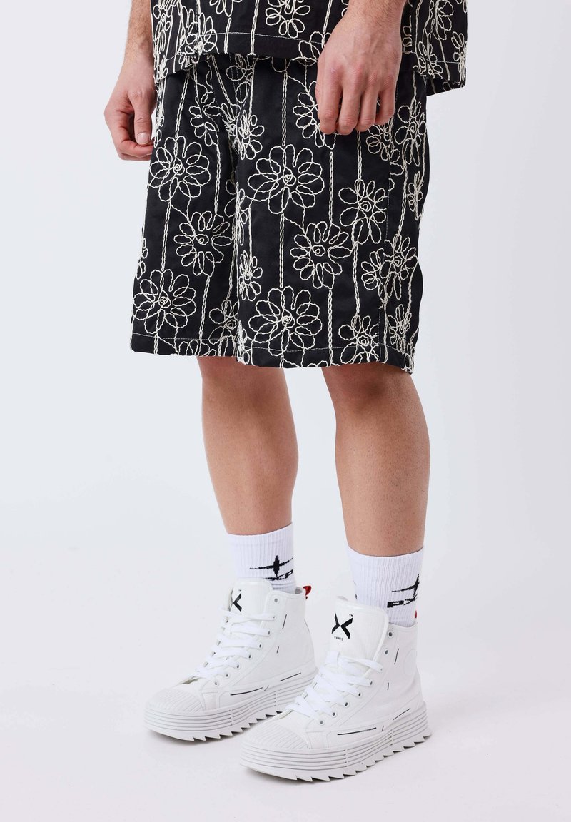 Homme portant un short noir à imprimé floral blanc, des chaussettes blanches montantes avec un logo noir, et des baskets montantes blanches sur un fond uni.