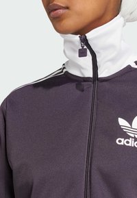 Chaqueta morada con cremallera y cuello blanco, que presenta tres rayas blancas, el logo de Adidas y una textura suave. La cremallera tiene un pequeño tirador con logo.