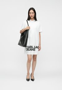 Sieviete, kura valkā baltu T-kreklu kleitu ar uzrakstu "KARL LAGERFELD JEANS", melnas augstpapēžu kurpes un nes lielu melnu pleca somu.