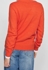 Pull en tricot orange avec un col ras du cou à côtes, des manches longues et une taille cintrée, porté avec un jean bleu. Texture et détails de couture visibles.