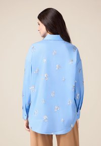 Helles blaues Bluse mit silberner Floralstickerei, langen Ärmeln mit Knopfmanschetten und einem leicht abgerundeten Saum auf der Rückseite.