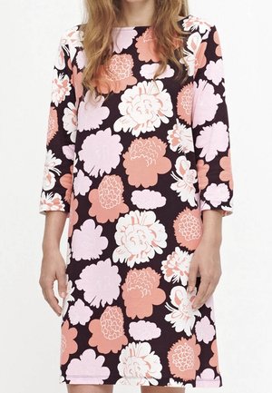 Femme portant une robe mi-longue à manches longues, ornée de grands motifs floraux roses et corail sur un fond sombre.