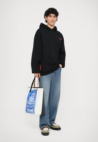 Schwarz-weißer Kapuzenpullover mit rotem Logo, hellblaue weite Jeans und eine weiße Tote-Bag mit blauem Grafikdesign. Schwarze Schuhe mit hellen Akzenten.