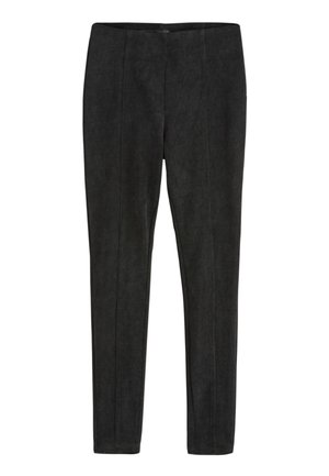 Junge Frau mit lockigem Haar steht an einer Straßenecke, sie trägt ein beiges kurzärmeliges Oberteil, schwarze Hose mit hoher Taille und schwarze Stiefeletten.