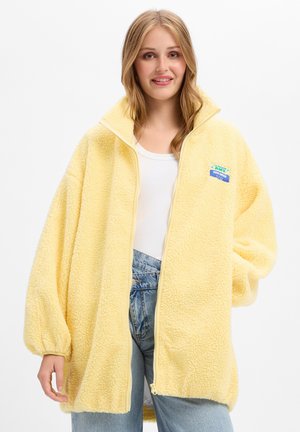 Jeune femme souriante, portant une veste polaire jaune oversize, un top blanc et un jean bleu sur un fond clair uni.