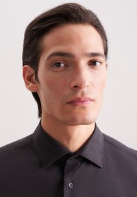 Una camicia nera con bottoni presenta un colletto strutturato e una texture liscia. Il modello ha capelli dritti e scuri e un'espressione neutra.
