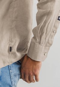 Helles beige Leinenhemd mit einem geknöpften Manschette und einem kleinen Etikett. Hand liegt auf blauen Jeans, eine Tasche ist sichtbar.