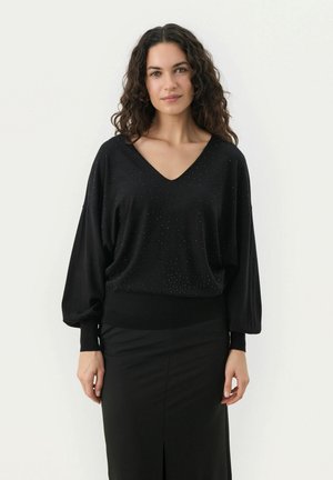 Pull noir avec décolleté en V, manches longues, et un motif texturé orné de strass dispersés. Associé à une jupe noire ajustée.