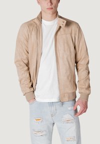 Giacca bomber in suede beige con chiusura a zip, polsini e colletto a costine, indossata sopra una t-shirt bianca e jeans chiari strappati.
