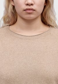 Νεαρό άτομο με ανοιχτό ξανθά μαλλιά που φοράει μπεζ πλεκτό πουλόβερ, εμφανίζεται από τον λαιμό μέχρι το στήθος με απλό φόντο.