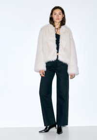 Jachetă albă din blană artificială cu textură pufoasă, asortată cu un top negru mulat și blugi din denim închis, cu talie înaltă și cusături laterale cu ținte. Tocuri negre ascuțite.