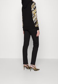 Černá mikina s vzorovanými rukávy, černé kalhoty a černé a zlaté podpatky se slingback designem a textovými detaily na popruzích.