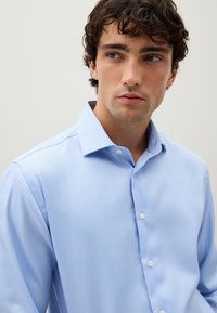 Giovane uomo con capelli ricci e scuri che indossa una camicia a collare azzurro chiaro, che guarda leggermente verso destra su uno sfondo semplice.