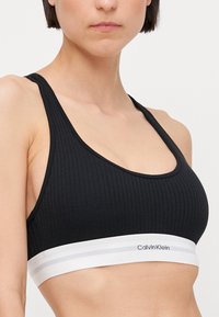 Fekete bordázott sportmelltartó széles fehér pánttal, amelyen a Calvin Klein logo látható. Simább textúra, U-alakú dekoltázs és versenyhát kialakítás.