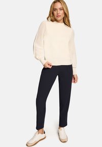 Femme debout portant un pull en maille crème avec des manches texturées, un pantalon fuselé bleu marine et des baskets blanches à semelles beige.