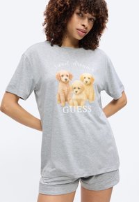 Femme portant un pyjama gris avec trois petits chiens et le texte "Sweet dreams" et "GUESS" sur le t-shirt, debout les mains sur les hanches.
