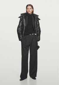 Black puffer jacket with a high collar, worn over a black turtleneck. Loose black trousers with a belt. Smooth textures and sleek design.