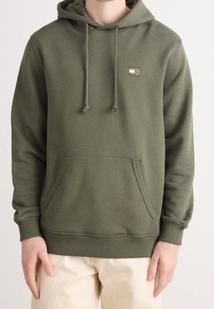 Personne portant un sweat à capuche vert olive avec poche avant et logo "Tommy Jeans" sur la poitrine gauche, associé à un pantalon beige clair.