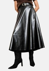 Femeie îmbrăcată cu o fustă A-line, lucioasă, cu model de crocodil, de culoare neagră, cu o curea și cizme cu toc din suede negru.