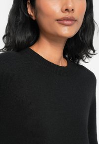 Suéter preto de malha com decote redondo, punhos canelados e uma textura suave, parcialmente exibido na parte superior do corpo de um modelo.