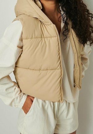 Woman wearing beige sleeveless puffer vest over cream long-sleeve shirt and shorts, with hand in pocket and curly hair visible.