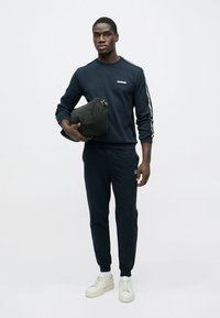 Sweatshirt azul-marinho com riscas laterais brancas e logótipo, combinado com calças de treino a condizer. Modelo segura uma mala preta, calçando sapatilhas brancas.