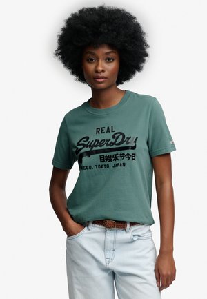 Kvinde med krøllet hår iført grøn "SuperDry" t-shirt og lyseblå jeans, poserer med den ene hånd i lommen mod hvid baggrund.
