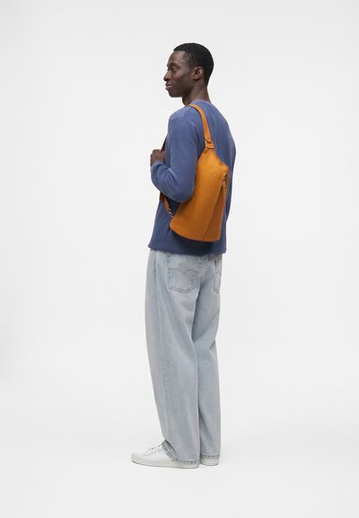 Jeune homme portant un pull bleu, un jean clair et large, des baskets blanches et un sac orange en bandoulière, regardant sur le côté.