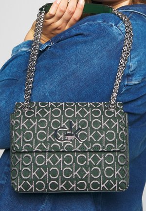 Zelená kabelka CK se vzorem a stříbrným řetězovým popruhem, kterou drží osoba v modré denimové bundě na jednobarevném pozadí.