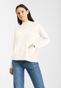 Jasno beżowy sweter dzianinowy z wysokim dekoltem, prążkowanym wykończeniem i dwiema kieszeniami z przodu. Noszony z niebieskimi dżinsami.