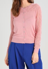 Ljusrosa cardigan med en texturerad, mönstrad stickad övre del, prydd med sex dekorativa knappar och ribbad manschetter, i kombination med blå byxor.