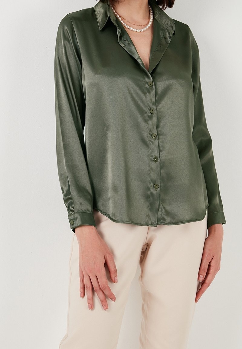 Satin green long-sleeve shirt with a pointed collar, front button closure, and cuffs, paired with beige pants. Smooth texture and glossy finish.