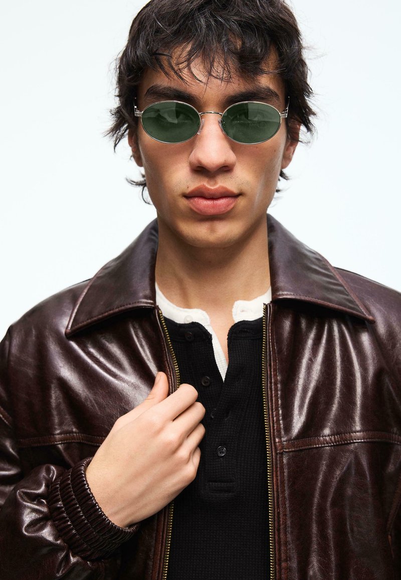 Jeune homme portant des lunettes de soleil rondes vertes et une veste en cuir marron foncé par-dessus une chemise noire boutonnée, tenant le col de la veste.