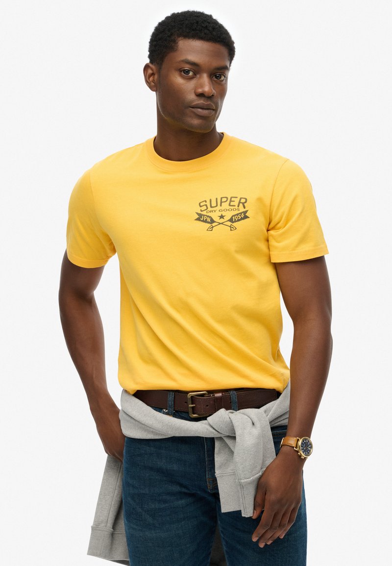 Camiseta de algodón amarilla con impresión y gráfico en negro que dice "SUPER DRY GOODS". Combinada con jeans de color azul oscuro y una sudadera gris atada a la cintura.