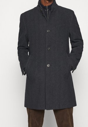 Homme portant un manteau en laine gris foncé boutonné, les mains dans les poches, et un pantalon marron, sur un fond clair uni.