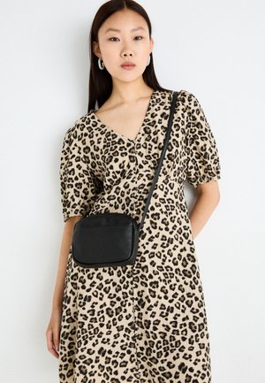 Žena v béžových šatech s leopardím vzorem, s nadýchanými rukávy a černou crossbody kabelkou, stojící na bílém pozadí.
