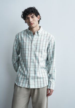 Νεαρός άνδρας με σγουρά μαλλιά, που φοράει ελαφρύ καρό πουκάμισο με κουμπιά και μπεζ παντελόνι, στέκεται μπροστά σε απλό ανοιχτό γκρι φόντο.