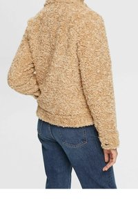Manteau beige, duveteux avec une finition texturée, doté d'un col et de manches longues, associé à un jean bleu. Aucun élément métallique ou accent visible.