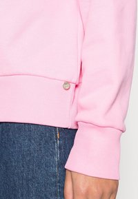 Sudadera de color rosa claro con puños y dobladillo acanalados, con un pequeño detalle de logo metálico en el lateral. Combinada con vaqueros oscuros.
